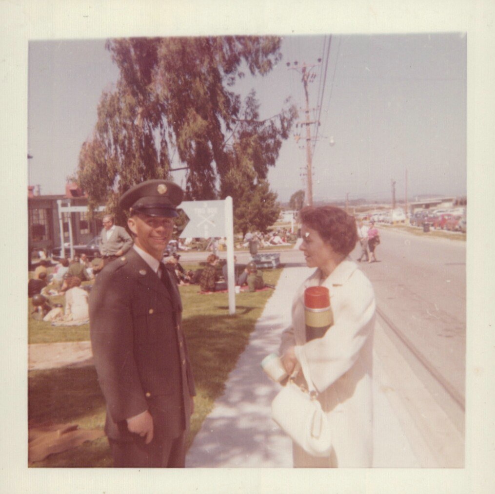 A smiling man in a U.S. Army dress uniform stands beside a woman in a white coat on a sunny day at what appears to be a military base, with groups of people relaxing on the grass behind them.