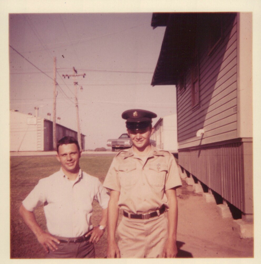 Two young men — one in civilian clothes, one in a U.S. Army tan uniform and peaked cap — stand together outside barracks-style buildings on what appears to be a military base, circa 1960s.