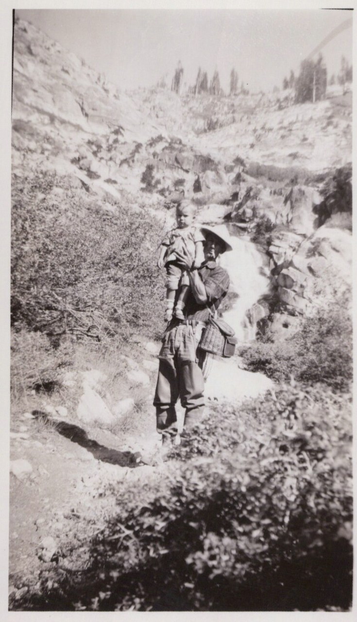 An adult in a wide-brimmed hat carries a small child on their shoulders while standing on a rocky trail, with a waterfall and coniferous trees visible in the background.