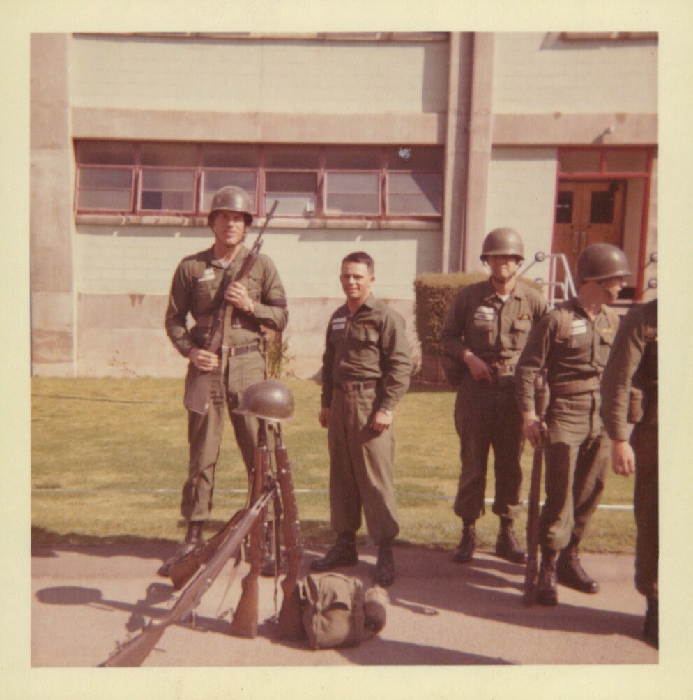 Group of soldiers in olive-drab uniforms and M1 helmets standing in front of an institutional building, with stacked rifles and a helmet at their feet, circa 1960s.