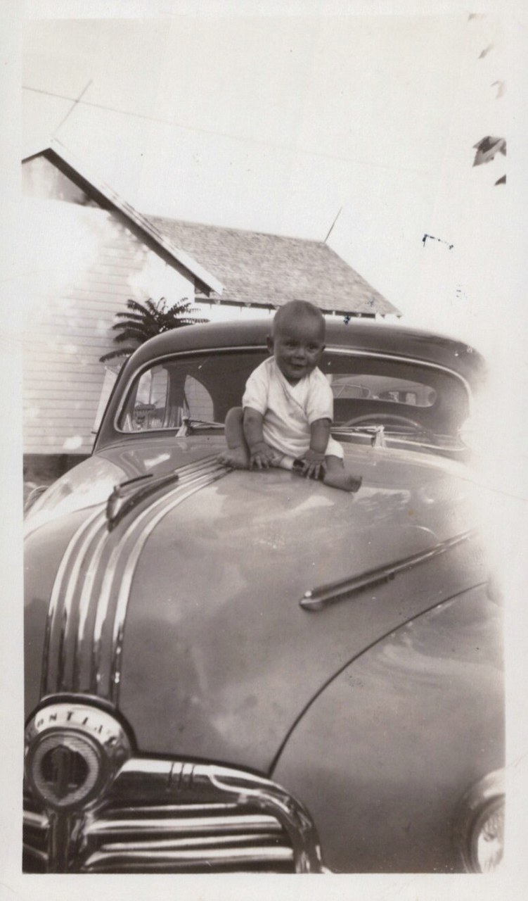 A baby in a white onesie sits on the hood of a vintage Pontiac automobile, parked in front of residential buildings; black-and-white snapshot, circa late 1940s–early 1950s.