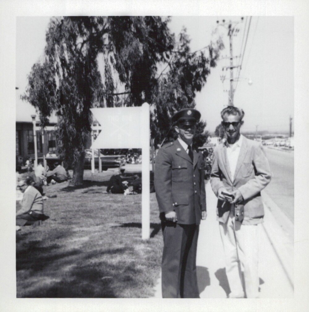 Two men — one in U.S. military dress uniform and one in civilian clothes — stand on a sunny walkway near a grass lawn, with a large tree and low buildings in the background; black-and-white snapshot.