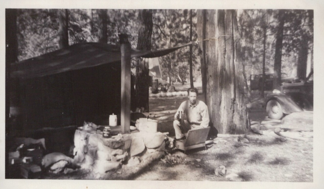 A man sits in a folding camp chair beside a large tree at a rustic campsite under a canvas tarp, surrounded by camp supplies and a vintage car, in a conifer forest