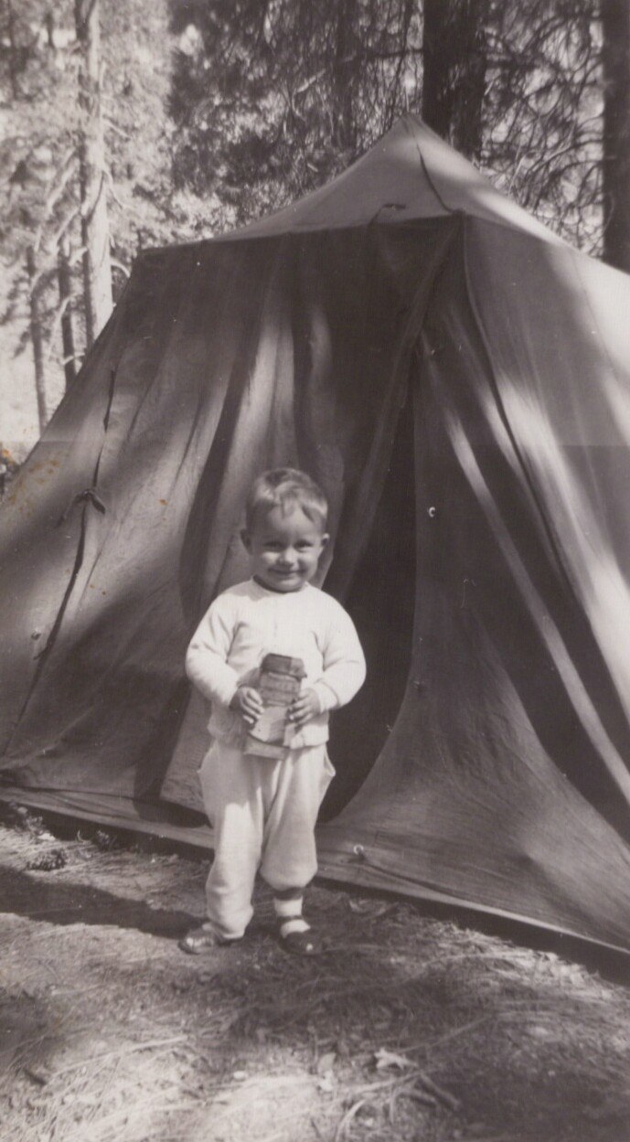 A smiling young child in light-colored clothing holds a small jar while standing in front of a large canvas camping tent surrounded by pine trees, in a black-and-white photograph.