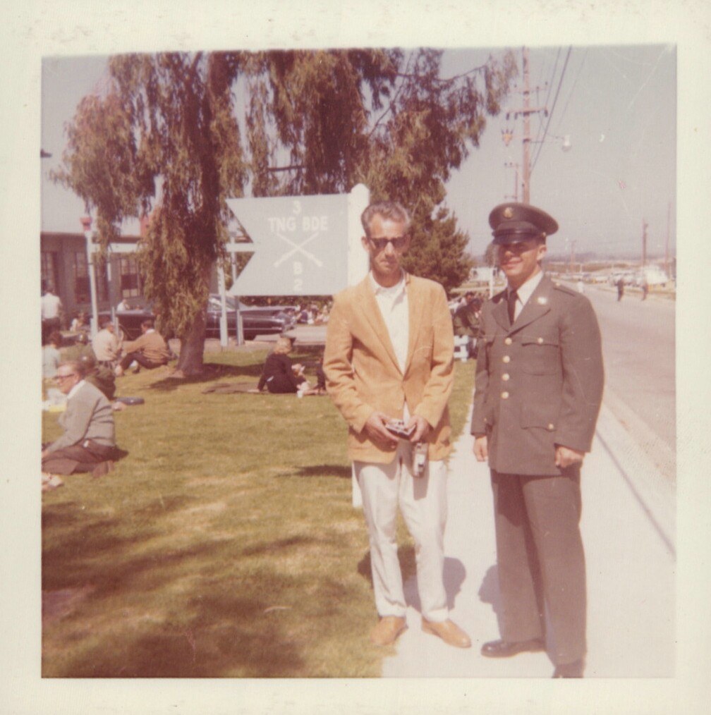 Two men — one in civilian clothes and one in a U.S. Army dress uniform — stand together in front of a Training Brigade sign on a military base, with people relaxing on the lawn behind them.
