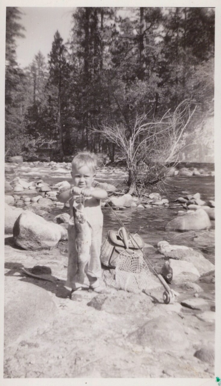 A toddler in overalls stands on a rocky riverbank holding a large fish, with a fishing creel, net, and rod nearby, tall pine trees and a mountain river in the background.
