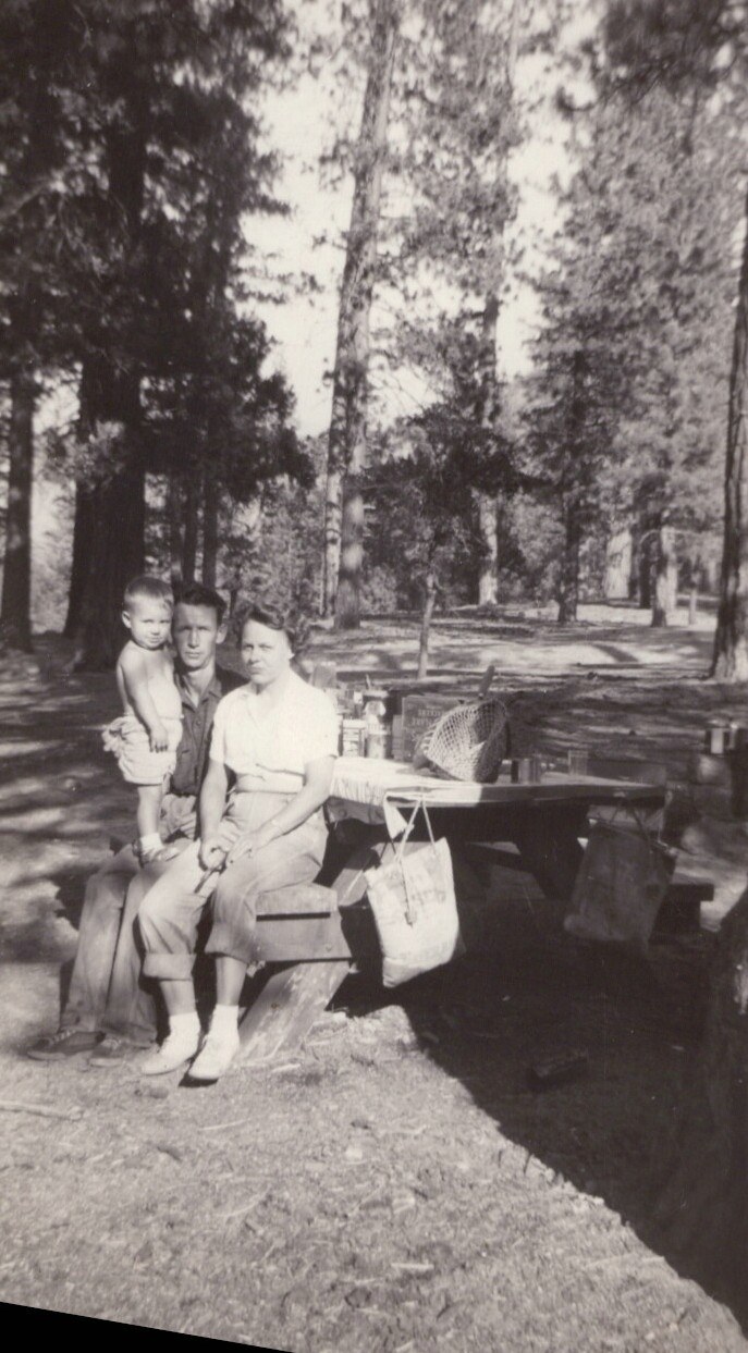 Black-and-white photo of two adults and a small child at a wooded picnic area, seated and standing at a wooden picnic table surrounded by tall conifers