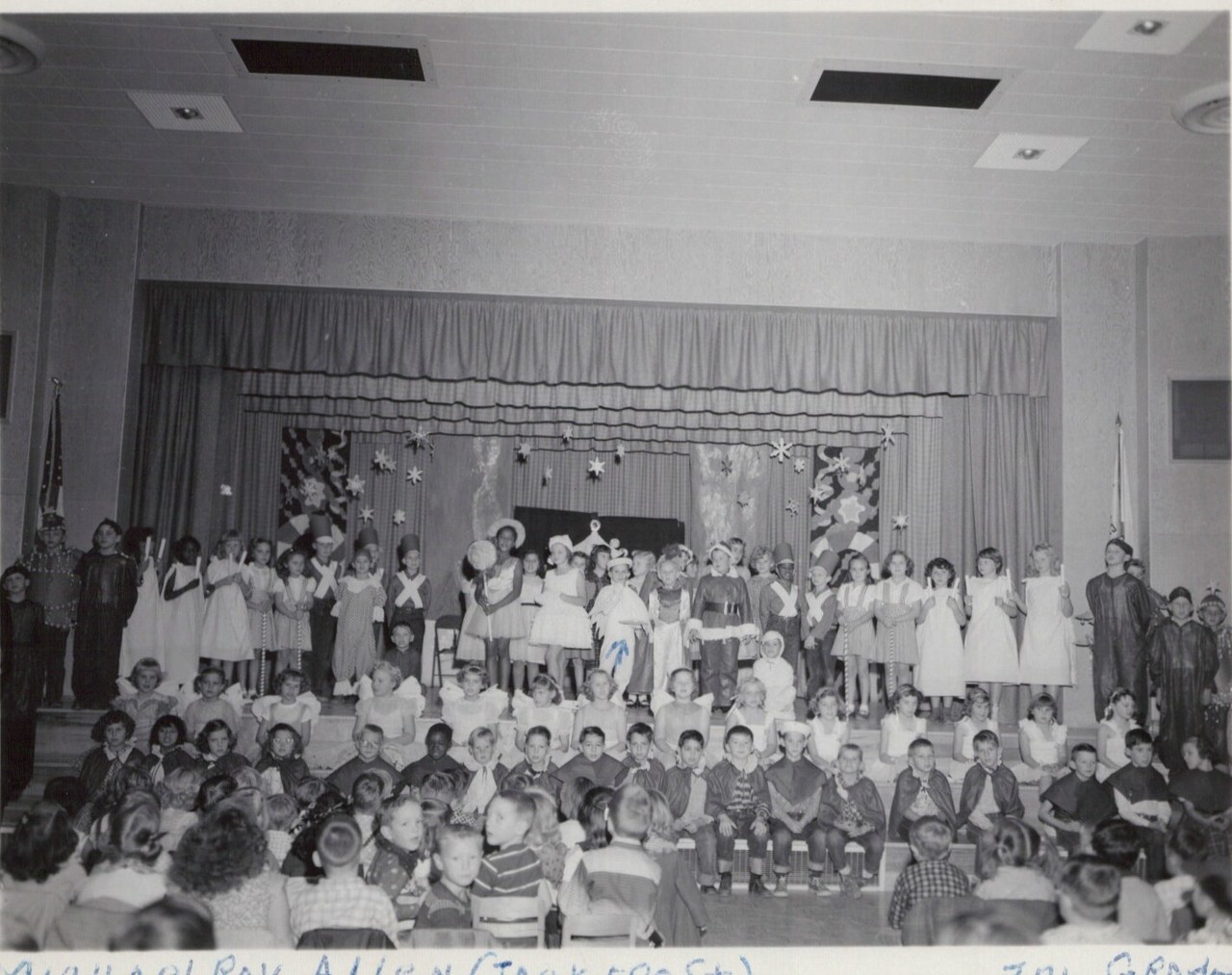 Black-and-white photo of a large group of costumed elementary school children performing a holiday pageant on a decorated school auditorium stage, with an audience of young children seated in the foreground