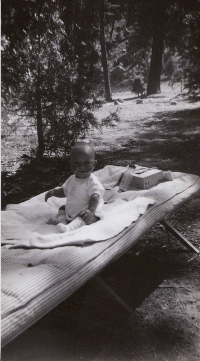 A baby in a white onesie sits on a blanket over a folding camp cot outdoors in a sunny, wooded setting, black-and-white photo