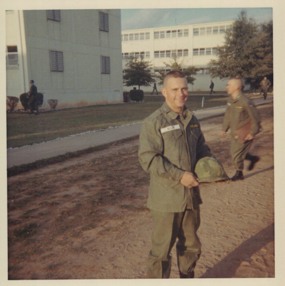 A smiling U.S. Army soldier in olive-drab fatigues holds a steel helmet on a military base, with large institutional buildings and other soldiers visible in the background.