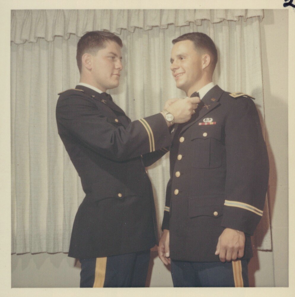 Two young men in U.S. Army dress uniforms, one pinning an insignia onto the other's uniform, standing before white curtains.