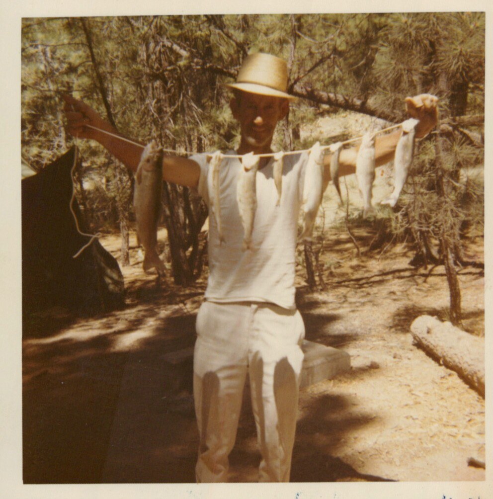 A person in a straw hat and white clothes stands outdoors at a wooded campsite, arms outstretched, holding a stringer of small fish.