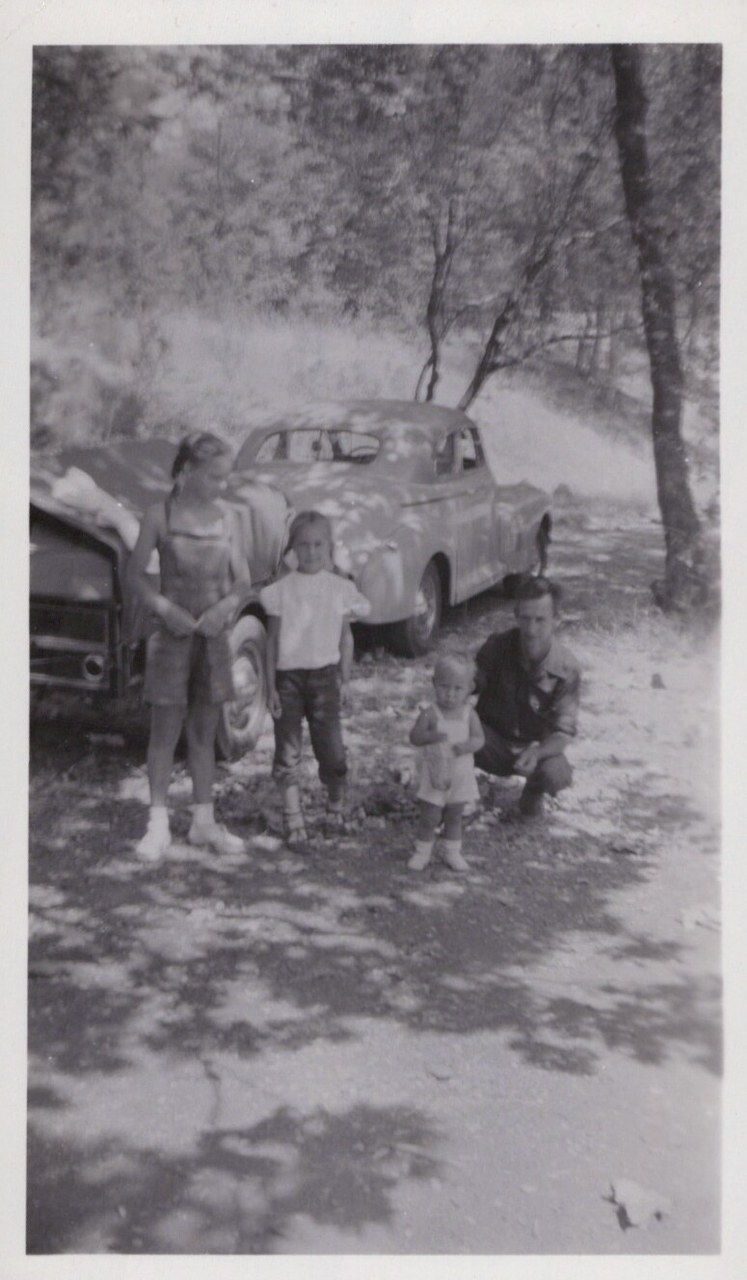 Black-and-white snapshot of four children and/or young people standing outdoors near two vintage cars parked among trees on a dirt surface, circa late 1940s–mid 1950s.