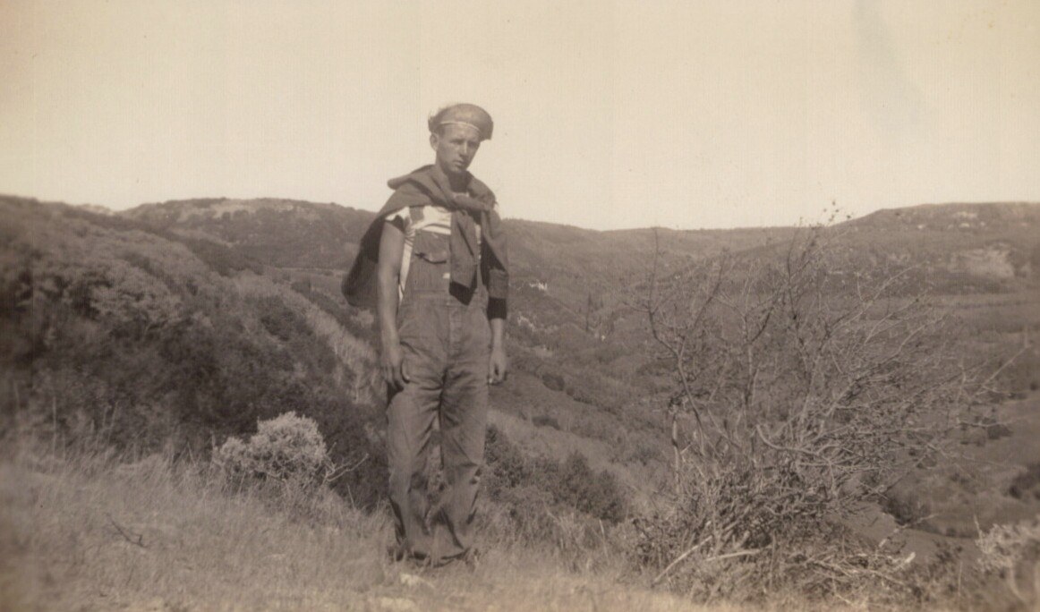 A young man in denim overalls and a flat cap stands on a scrubby hillside, with rolling brush-covered hills in the background, in a sepia-toned vintage photograph.