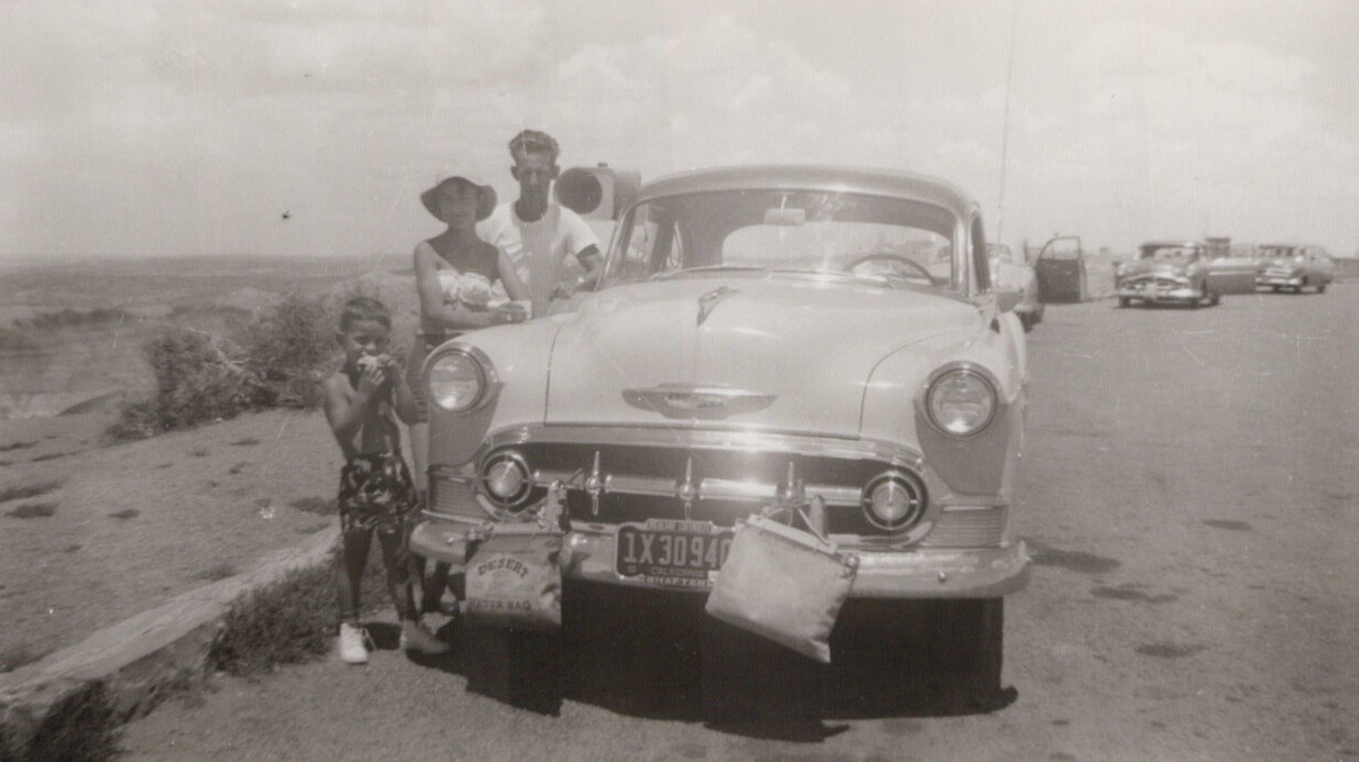 Black-and-white 1950s photo of two adults and a child standing in front of a large American sedan in an arid desert parking area, with a canvas desert water bag hanging from the front bumper.