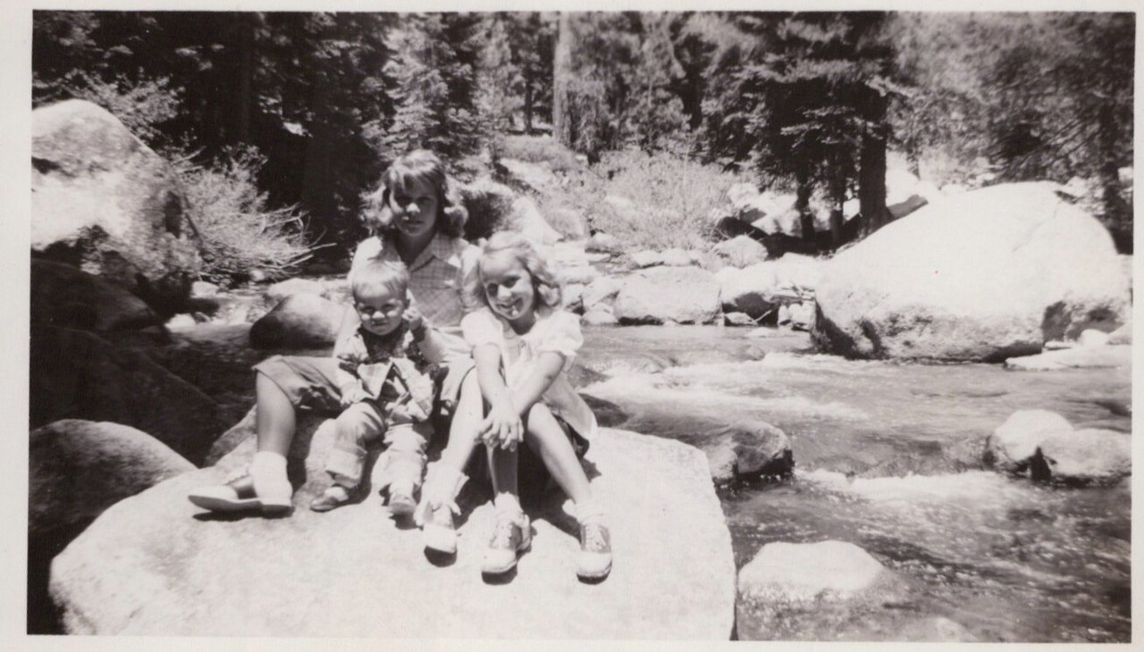 Three children — an older girl, a toddler, and a younger girl — sitting together on a large granite boulder beside a rocky mountain stream, surrounded by conifers, in a black-and-white photograph.