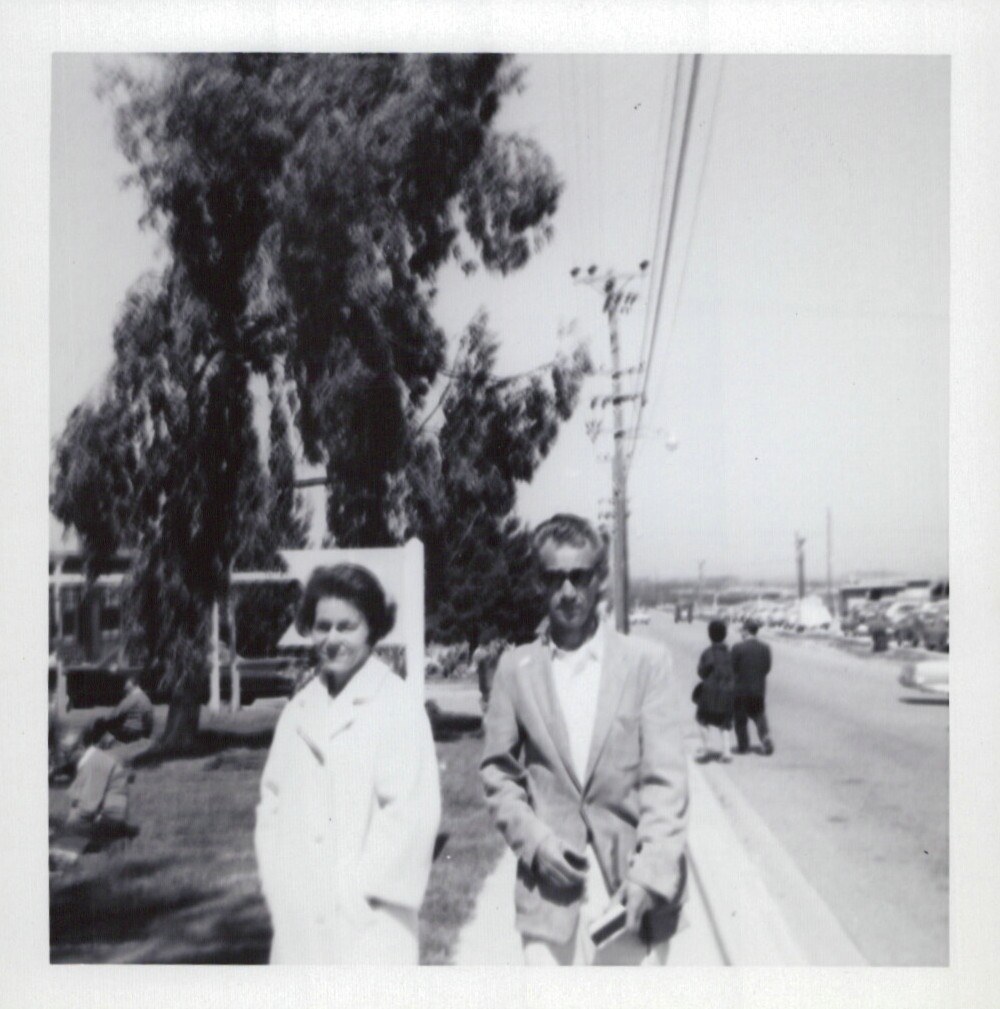 A man and woman walk along a sidewalk in a public outdoor setting, surrounded by large trees and a wide road, in a black-and-white snapshot from approximately the late 1950s to mid-1960s.