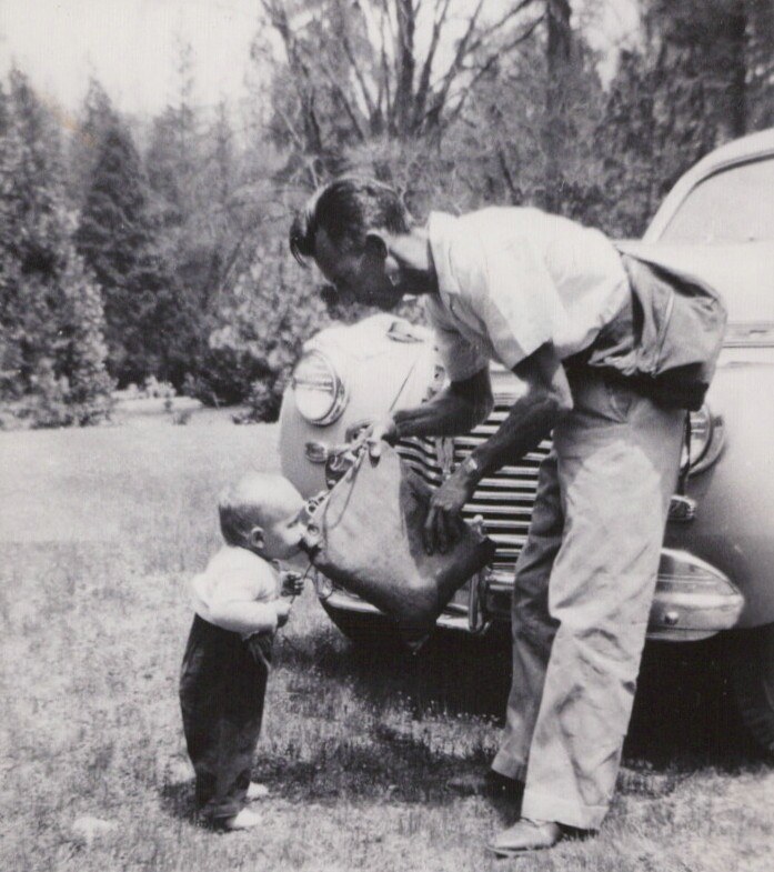 A young adult leans over the front of a vintage rounded car toward a toddler standing on a grass lawn, with trees in the background; black-and-white photo.