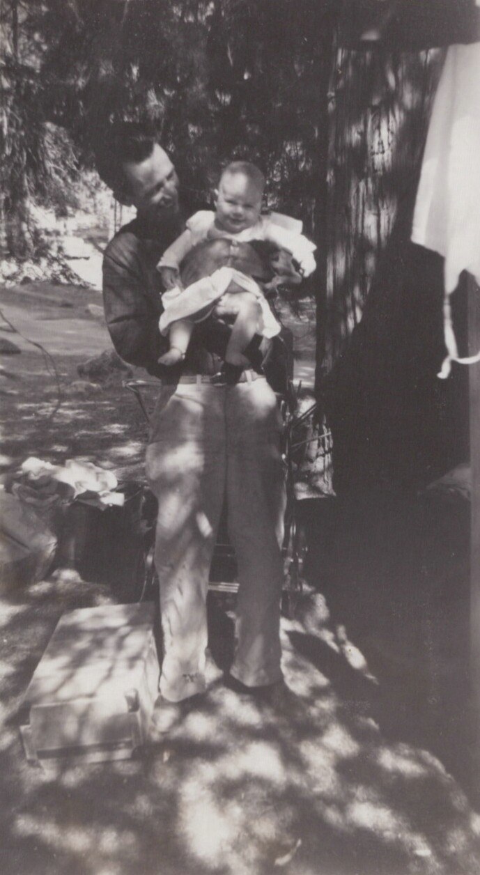 An adult holding a smiling infant outdoors in dappled sunlight, with a vintage pram visible in the background, black-and-white photograph