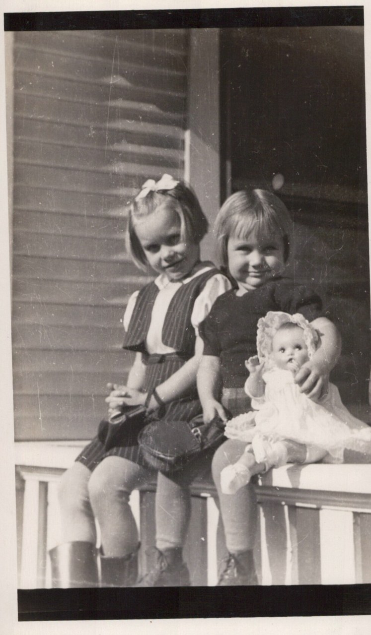 Two young girls sitting on a white wooden porch railing; one holds a baby doll dressed in lace, the other wears a large hair bow, black-and-white photo circa 1940s–1950s