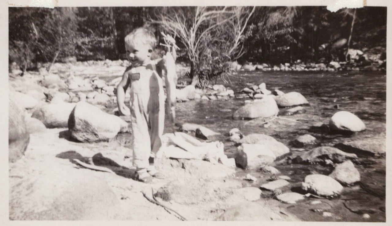 A young toddler in overalls stands on a rocky riverbank holding a twig, with a shallow rocky stream and trees in the background, in a vintage black-and-white photo.