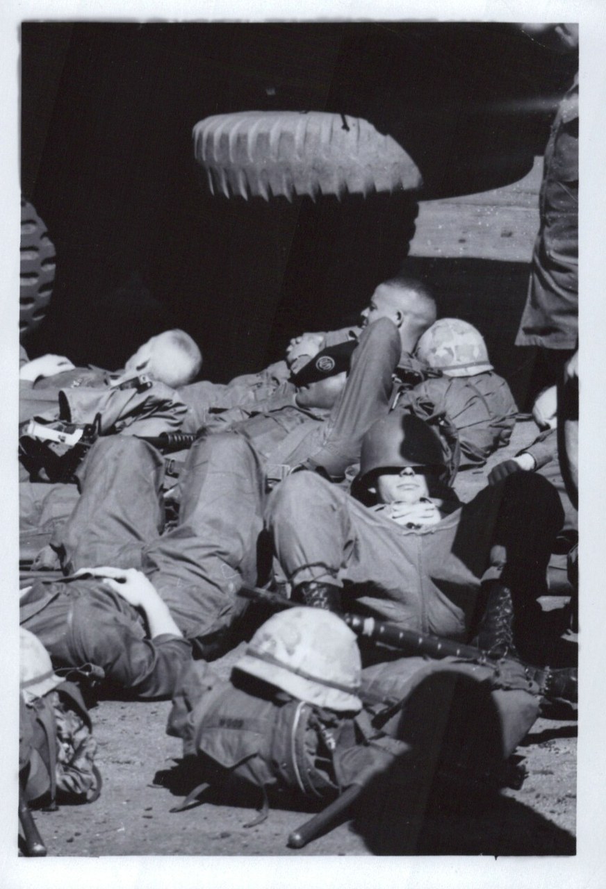 Black-and-white photo of several soldiers in combat fatigues and helmets resting on the ground beside a large military vehicle, carrying rifles and web gear