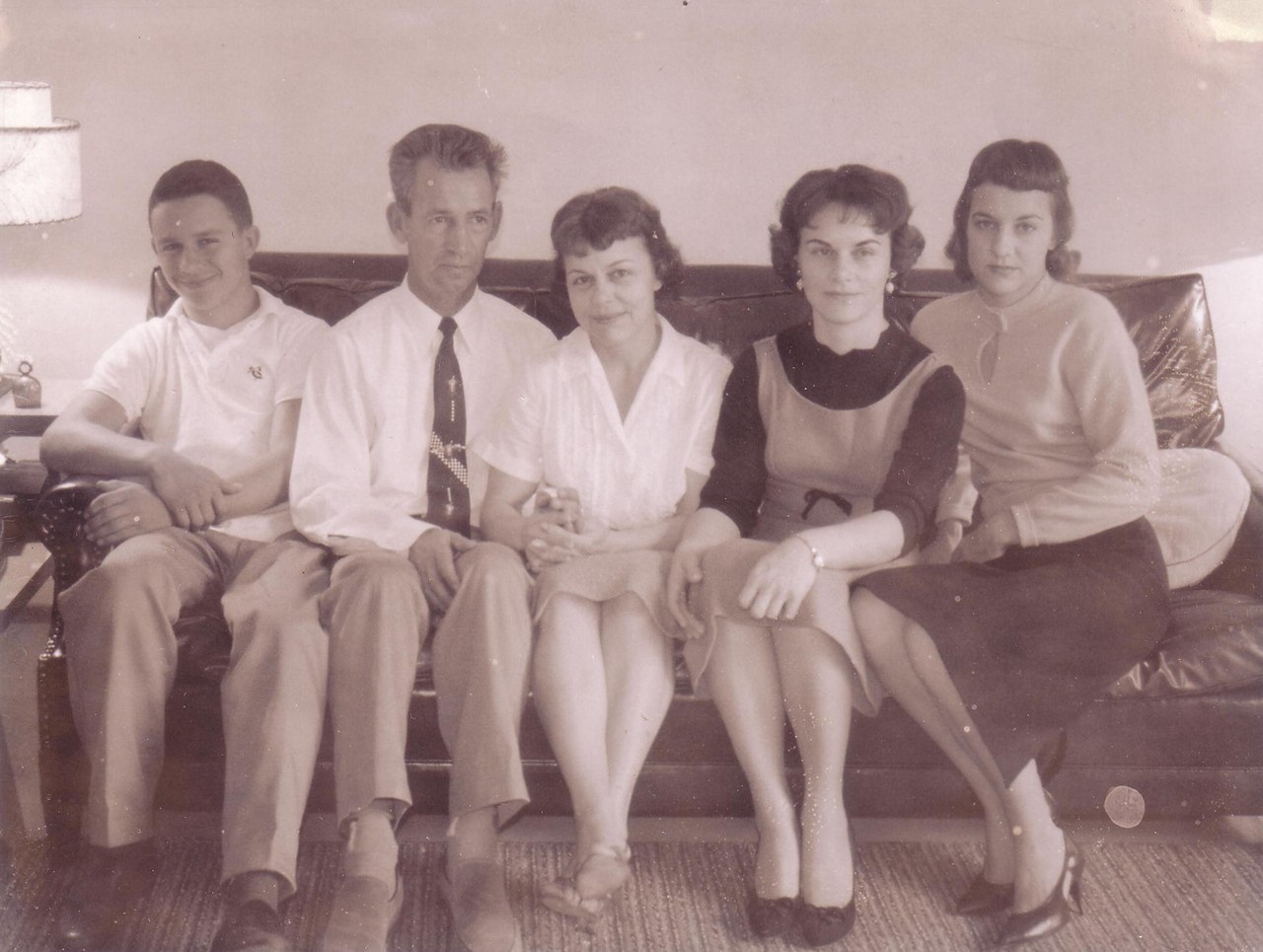 Five people — one boy, one adult man, and three women or girls — seated together on a dark leather sofa in a home living room, posed for a formal portrait, in a sepia-toned vintage photograph.