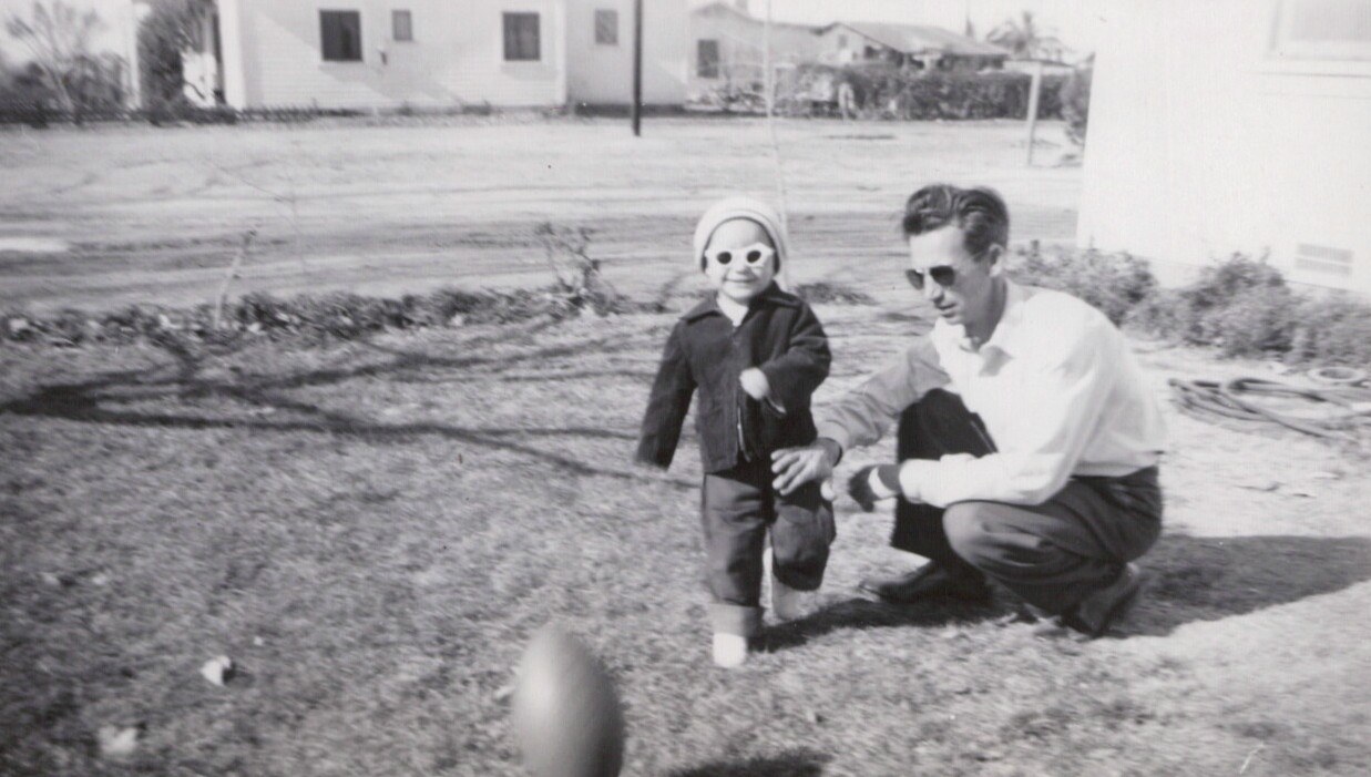 Black-and-white photo of a toddler in a jacket, hat, and novelty sunglasses walking on a lawn toward a crouching adult man in sunglasses, with a ball blurred in the foreground and residential buildings in the background.