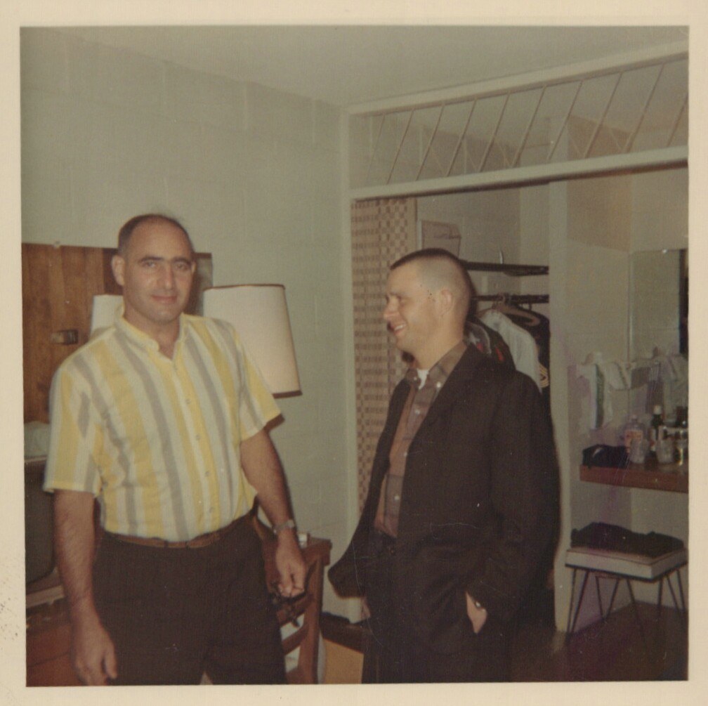 Two smiling men standing in a bedroom with an open closet, lamp, and mid-century furnishings, likely photographed in the 1960s.