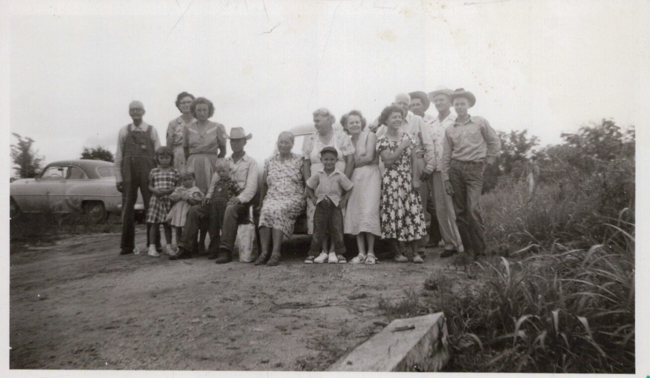 Large group of men, women, and children posed outdoors near a vintage car in a rural setting, black-and-white photo circa late 1940s–mid-1950s