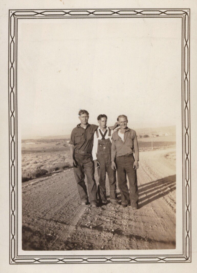 Three men of different ages standing together on a rural dirt road in an arid landscape, sepia-toned photo with decorative border, likely 1930s–1940s American West