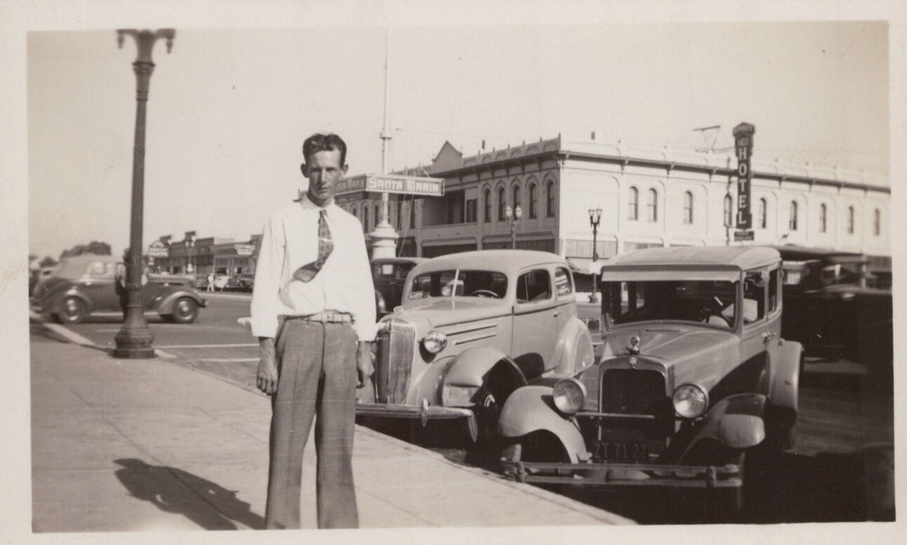 A man in a shirt and tie stands on a sunny sidewalk in downtown Santa Maria, California, with 1930s-era parked cars and a hotel building behind him.