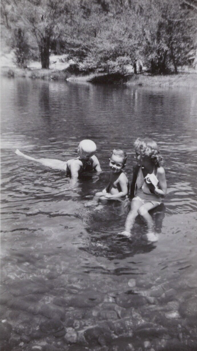 Three young children wading and playing in a shallow, rocky creek, black-and-white photo, likely mid-20th century.