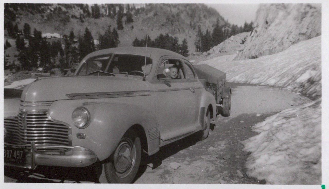 A vintage early-1940s American coupe with license plate B87 497 on a narrow snow-walled mountain road; a young child leans out the passenger window, with a canvas-topped truck behind and rocky forested peaks in the background.