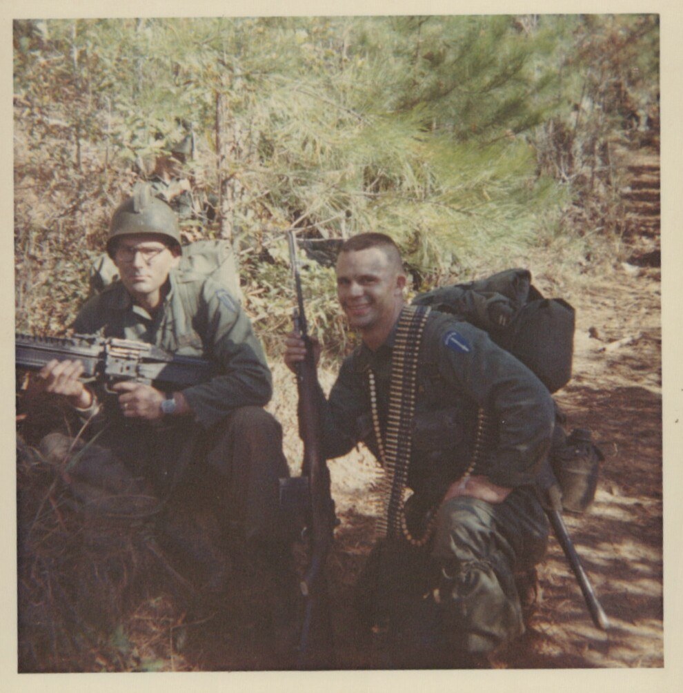 Two U.S. soldiers in combat gear with rifles and ammunition belts posing in a wooded area, likely during the Vietnam War era