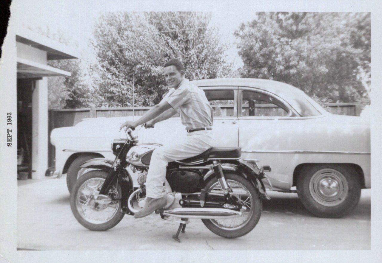 Man in light clothing posing on motorcycle with car behind in driveway.