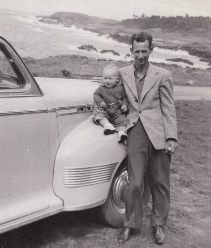 A man in a sport coat stands beside a vintage American car at a coastal overlook, steadying a toddler seated on the front fender, in a black-and-white photo from the late 1940s or early 1950s.