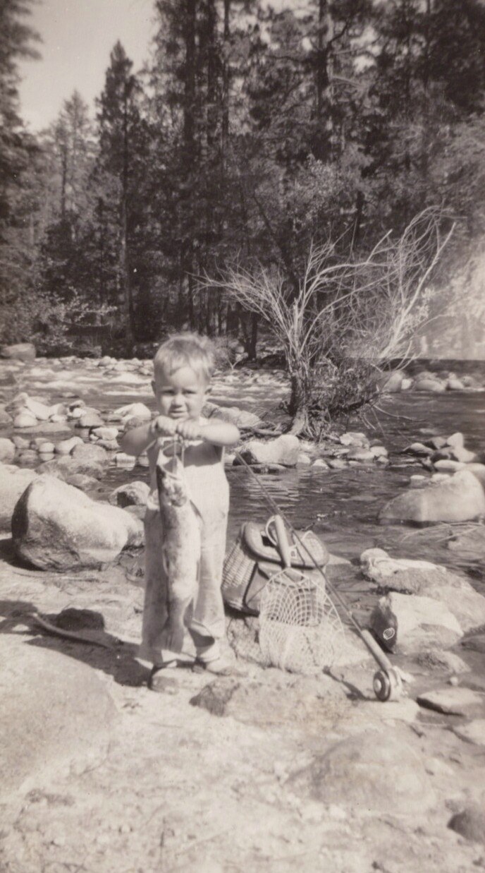 A young toddler in overalls proudly holds up a caught fish on a rocky riverbank, with a wicker creel and fishing net nearby and tall pine trees in the background.
