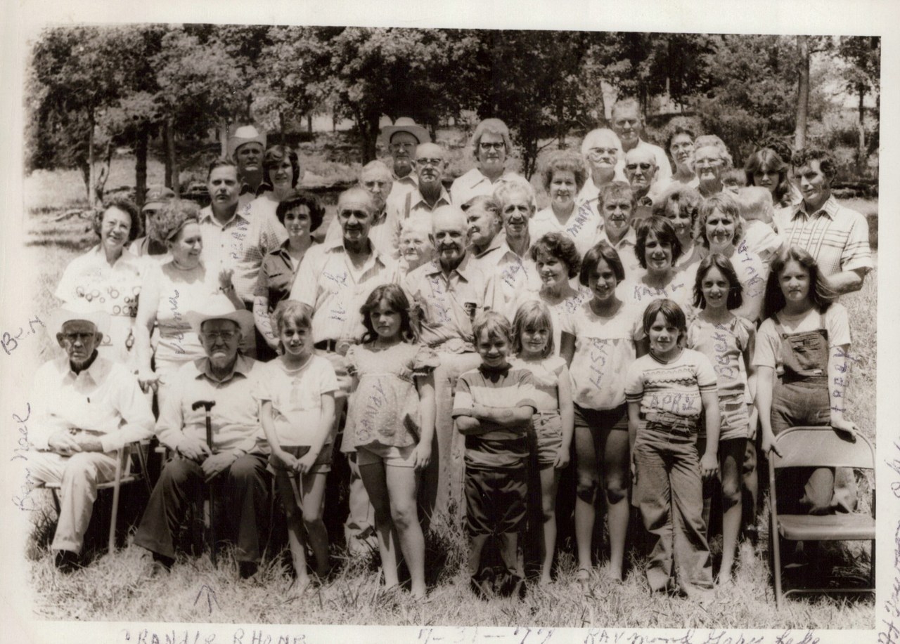 Black-and-white group portrait of approximately 30–35 people of multiple generations posed outdoors on grass with trees behind them; handwritten names annotate the photo; two elderly men sit in folding chairs at the front.