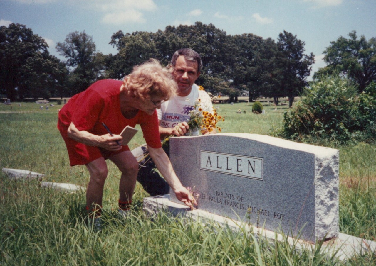A woman in red and a man in a white athletic shirt kneel at an "ALLEN" family granite headstone in a green cemetery, with flowers placed on the monument.