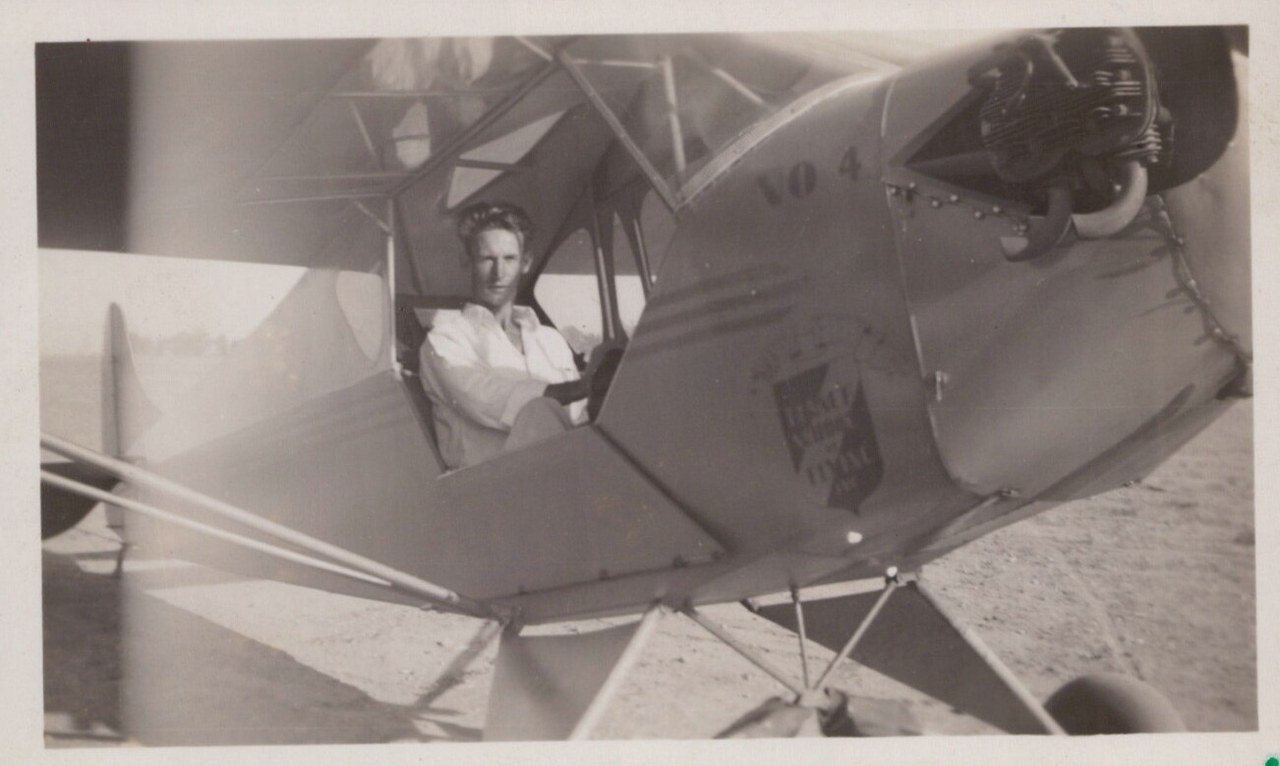 A young person sitting in the open cockpit of a vintage fabric-covered light aircraft on an unpaved airfield, black-and-white photo circa 1930s–1940s.