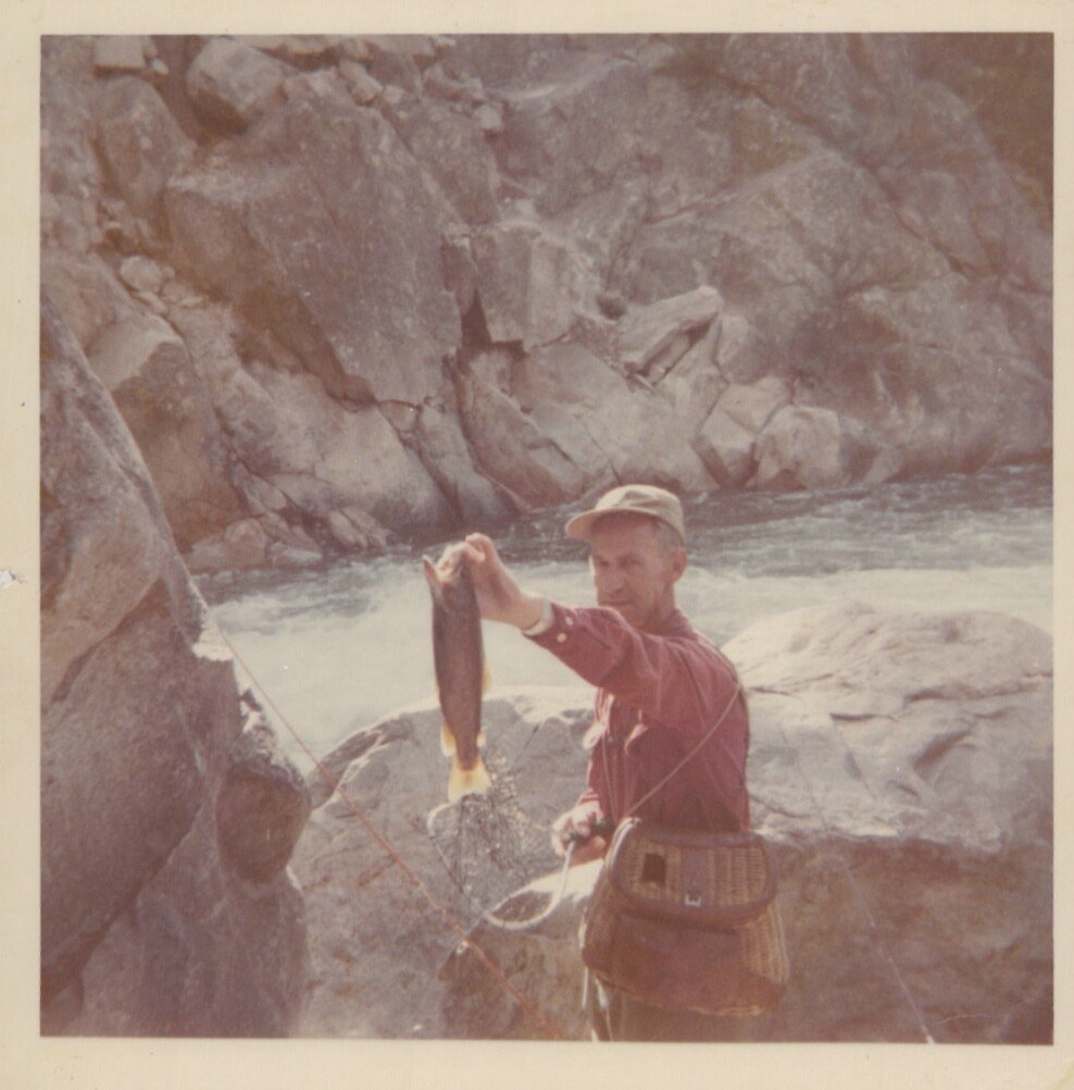 A man in a red shirt and cap holds up a freshly caught fish beside a rocky mountain river, with a wicker creel at his waist and a fishing net in hand.