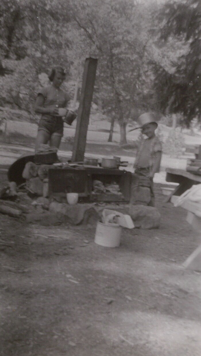 Black-and-white photo of an adult woman and a child in a wide-brimmed hat cooking at a large circular outdoor camp stove with a tall flue pipe, in a wooded campground setting.