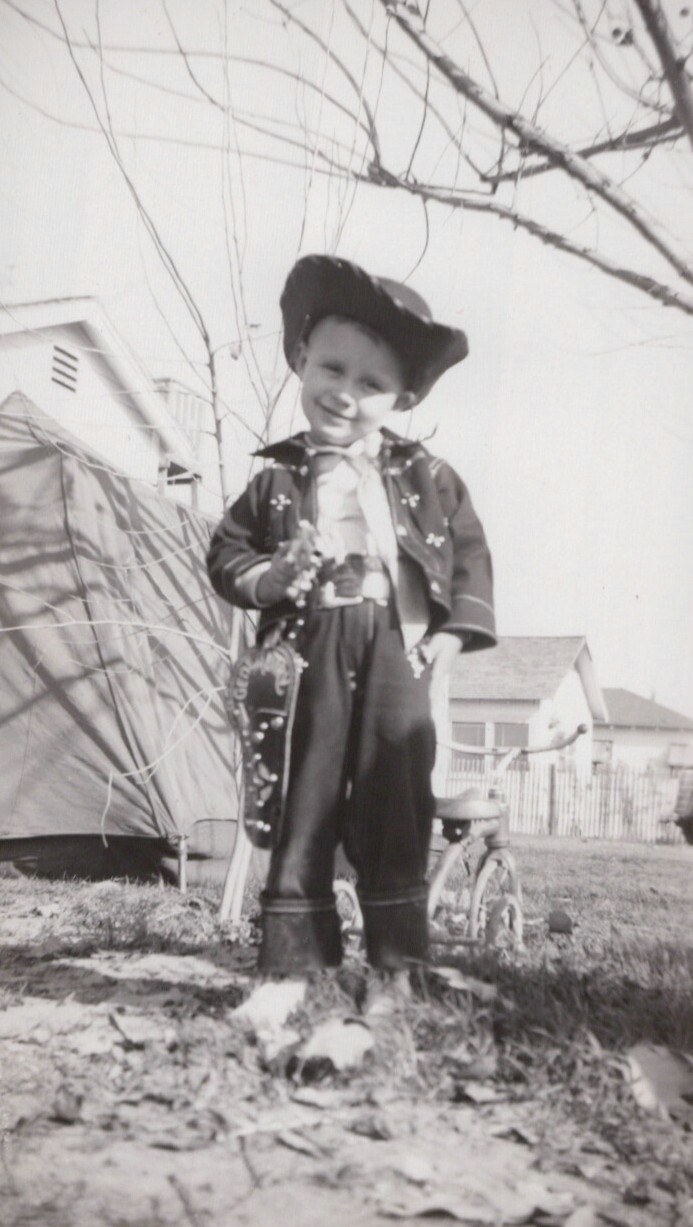 Young child in cowboy outfit standing outdoors near bare tree.