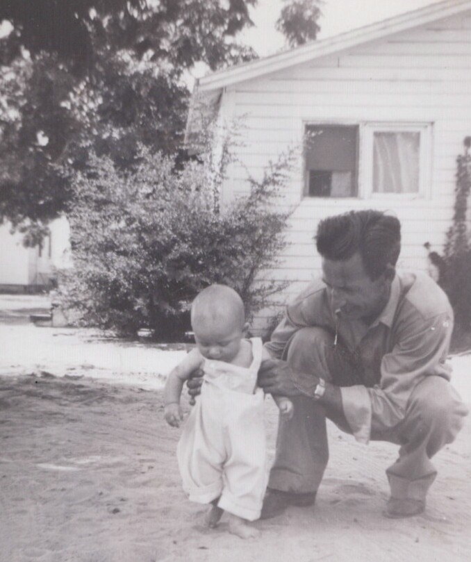 A man in a light work jacket crouches behind a barefoot toddler in bib overalls on sandy ground, helping the child stand, in front of a white clapboard house — black-and-white photo.