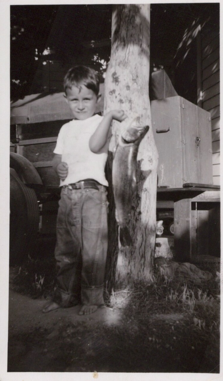 A barefoot young boy in a white T-shirt and jeans holds a large fish against a tree trunk in a rural farmyard, black-and-white photo